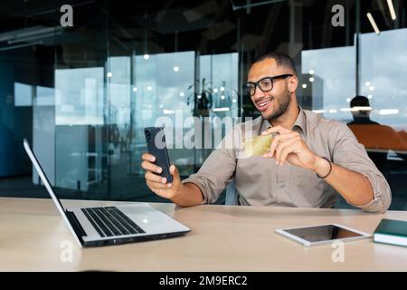 Uomo d'affari ispanico di successo all'interno dell'ufficio, uomo in camicia sorridente e felice tenendo la carta di credito e il telefono della banca, facendo il trasferimento di denaro e lo shopping in linea. Foto Stock