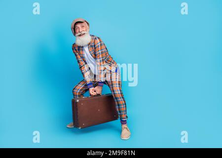 Foto a tutta lunghezza di uomo allegro pensionato indossare costume a scacchi sollevamento pesante bagaglio vuoto spazio isolato di colore blu sfondo Foto Stock