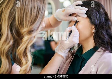 Primo piano di makeup femminile artista in guanti sterili applicazione matita palpebra superiore cliente. Specialista di bellezza fare trucco professionale occhio su donna in salone di bellezza. Foto Stock