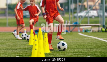 Esercizi di calcio dribbling per giovani giocatori. Bambini che corrono con palline da calcio trivello tra i coni gialli allenati Foto Stock