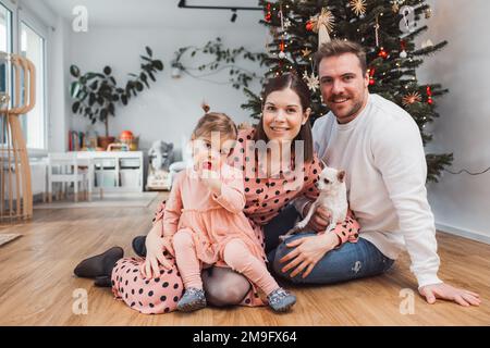 Bella giovane famiglia caucasica con bambina e chihuahua seduti sul pavimento accanto all'albero di Natale Foto Stock