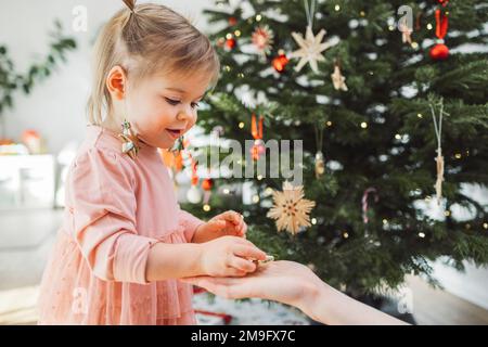 Bambina in abito rosa con ornamenti di Natale appesi fuori le orecchie, prendendo un altro ornamento dalla mano mamme Foto Stock