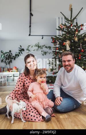 Ritratto verticale della giovane famiglia caucasica con bambina e chihuahua davanti all'albero di Natale Foto Stock