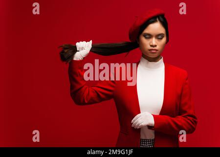 Modello asiatico in guanti e beretta capelli toccanti isolato sul rosso Foto Stock
