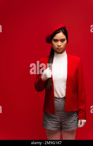 Elegante donna asiatica in guanti e giacca a contatto con i capelli isolato sul rosso Foto Stock