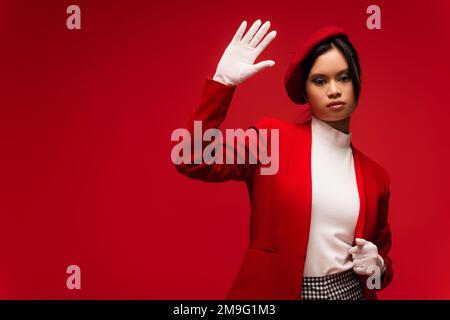 Donna asiatica in beret un guanto ondeggiante alla macchina fotografica isolato su rosso Foto Stock