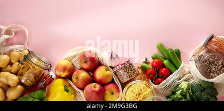 Vista dall'alto di appetitose mele fresche crude, patate, pomodori ciliegini, cetrioli, broccoli e peperone giallo in sacchi di cotone ecologici messi su rosa Foto Stock