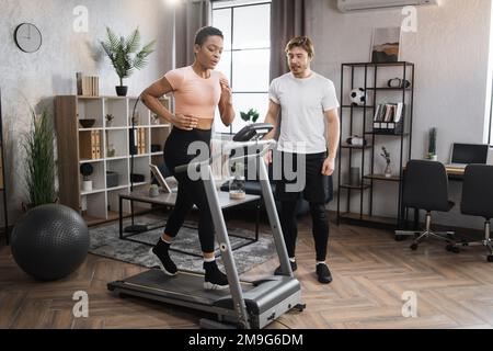 Home fitness allenamento sportivo donna e uomo allenamento sulla pista da corsa. Ritratto di una giovane donna africana focalizzata che indossa abbigliamento sportivo e tapis roulant e il suo allenatore uomo caucasico. Foto Stock