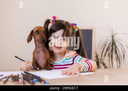 carino ragazza piccola disegna con il suo cane amico dachshund. Bambini e animali. Cani ammessi Foto Stock