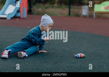 ragazzo con sindrome di down è seduto su una copertura di gomma sul parco giochi, raggiungendo per un giocattolo Foto Stock