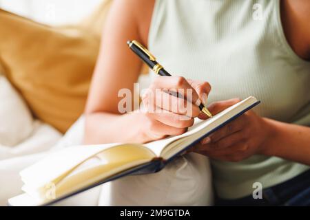 A metà della giovane donna biraciale che scrive in agenda mentre si siede sul divano a casa Foto Stock