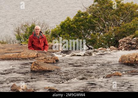 Uomo 60+ seduto su una roccia nella cascata EAS Fors, circondato da acqua, Mull, Scozia UK Foto Stock