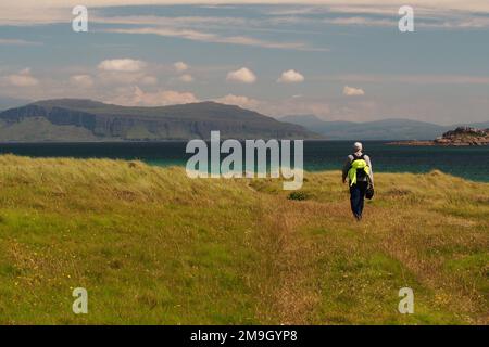 Uomo 60+ con zaino e cappotto ad alta visibilità, camminando attraverso l'erba su Iona verso il mare, The Sound of Iona, con il Mull in lontananza UK Foto Stock