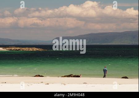 Un uomo che cammina sulla White Strand della spiaggia di Monk, Iona, Scozia UK, mostra la sabbia bianca e il mare blu profondo e le isole sullo sfondo Foto Stock