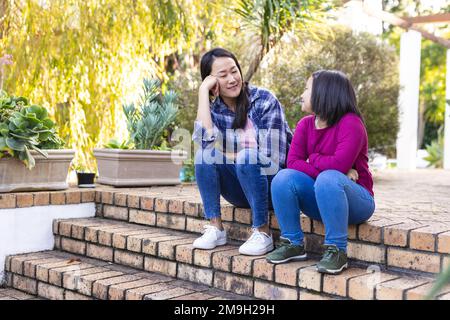 Felice madre asiatica e figlia che parlano e si siedono sulle scale in giardino Foto Stock