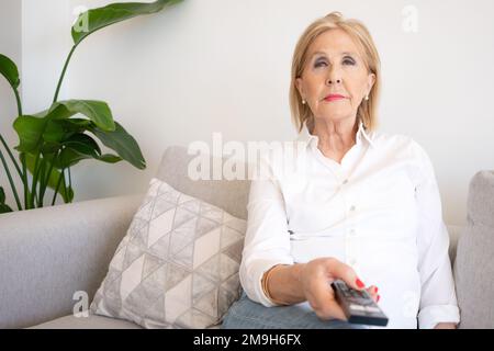 La donna anziana allegra ama la serata con una serie di spettacoli televisivi. Donna matura sorridente si siede sul divano con telecomando TV e commutazione dei canali Foto Stock