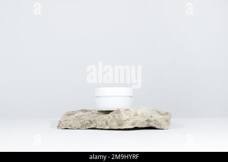 Un barattolo bianco di crema su un piedistallo da podio in pietra naturale, su un tavolo bianco, con ombre dure. Aspetto elegante del prodotto, identità. Foto Stock