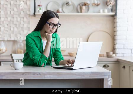 Focalizzato giovane donna designer, ingegnere, architetto che lavora da casa on-line. Seduti in cucina al tavolo, lavorando su un computer portatile. Tiene la testa con la mano, guarda il monitor. Foto Stock