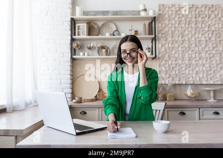 Una giovane studentessa, una freelancer studia, lavora online, a casa da un computer portatile. Si siede al tavolo, guarda la macchina fotografica, tiene gli occhiali, scrive in un taccuino, sorride. Foto Stock