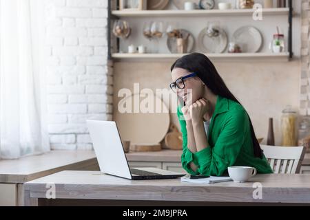 Una giovane donna felice in quarantena si siede a casa in cucina e comunica con amici, genitori, parenti attraverso una videochiamata da un portatile. Sorrisi, salumi. Foto Stock