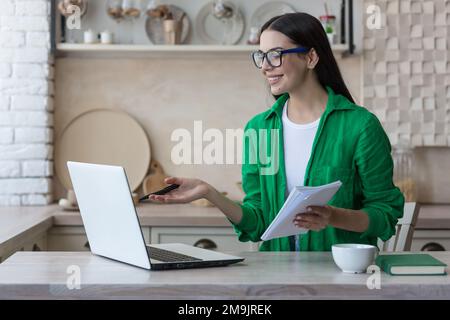 Una giovane studentessa, una freelancer studia, lavora online a casa da un computer portatile. Si siede al tavolo, ascolta la lezione, tiene un taccuino con una penna in mano, scrive, spiega. Foto Stock