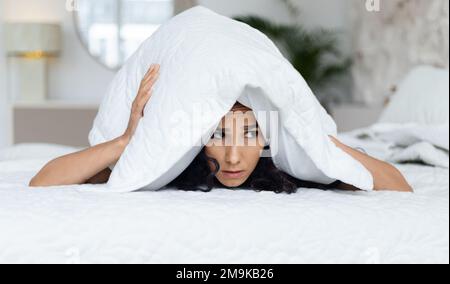 Triste musulmano millenario riccio femmina svegliato, si trova su letto bianco, copre la testa con cuscino, soffre di depressione Foto Stock