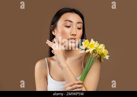 Giovane bella signora giapponese in posa con fiori di daffodil giallo isolato su sfondo marrone, studio shot Foto Stock