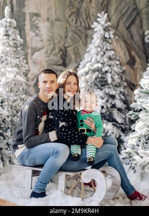 I genitori felici con il figlio del bambino posano sulla slitta in studio fra gli alberi di Natale decorati dalla neve artificiale. Foto Stock