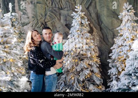 Genitori felici con bambino posa in studio tra alberi di Natale decorati da neve artificiale e ghirlande. Foto Stock