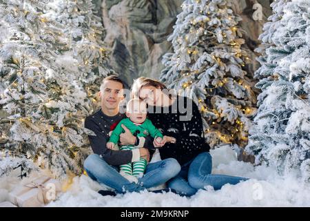 I genitori felici con il loro figlio del bambino posano seduti sul pavimento in studio fra gli alberi di Natale decorati dalla neve artificiale e dalle ghirlande incandescenti. Foto Stock