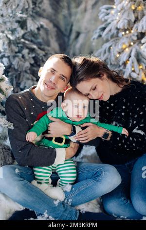 I genitori felici con il loro figlio del bambino posano seduti sul pavimento in studio fra gli alberi di Natale decorati dalla neve artificiale e dalle ghirlande incandescenti. Primo piano. Foto Stock