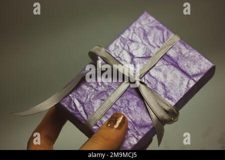 Scatola regalo viola con arco in seta d'argento in mano da donna su sfondo grigio. Regali. Shopping, shopping, regali ai cari. Regali in una scatola Foto Stock