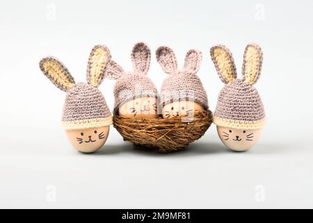 Quattro uova di Pasqua con volti divertenti e cappelli grigi uncinati con orecchie conigliate in un nido decorativo di sisal su sfondo grigio. Buon concetto di Pasqua. Gr Foto Stock