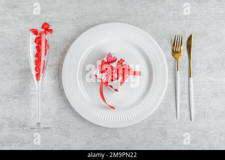 Cena di San Valentino con regalo su piatto bianco, posate e bicchiere di vino con cuori rossi su sfondo chiaro Foto Stock