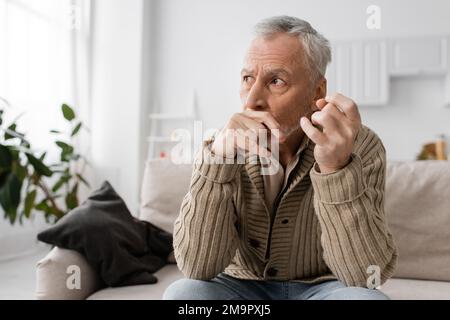 uomo depresso e ansioso con malattia di parkinson tenendo tremanti mani vicino al viso e guardando lontano a casa, immagine stock Foto Stock