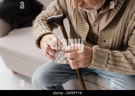 vista ritagliata dell'uomo anziano con il morbo di parkinson seduta con la canna da camminata e le pillole sul divano a casa, immagine di scorta Foto Stock