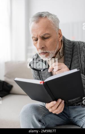 uomo anziano con sindrome di alzheimer che guarda nel notebook attraverso la lente di ingrandimento mentre si siede a casa, immagine stock Foto Stock