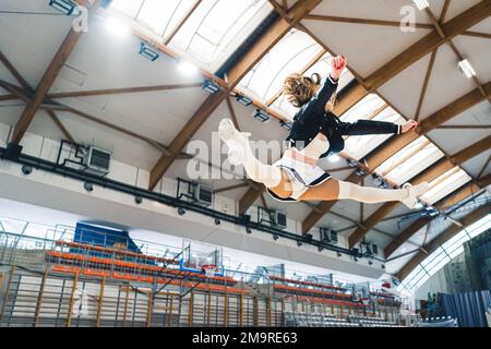 Vista laterale di un cheerleader nella sala sportiva che fa una spaccatura in aria. Il soffitto della sala sportiva e campo da basket sullo sfondo. Foto di alta qualità Foto Stock