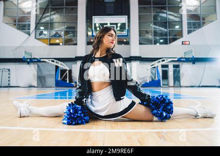 Giovane ragazza cheerleader in uniforme tenendo blu lucido pom e facendo una spaccatura in sala sportiva. . Foto di alta qualità Foto Stock