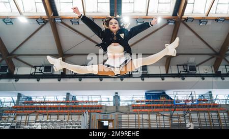 Cheerleader in uniforme bianca e nera nella sala sportiva facendo una spaccatura in aria. Il soffitto della sala sportiva sullo sfondo. Foto di alta qualità Foto Stock