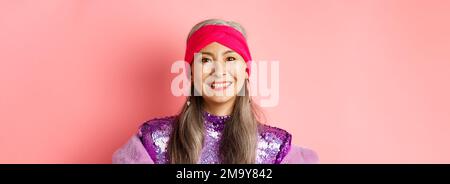 Primo piano della donna anziana asiatica alla moda con capelli grigi e un elegante abito da discoteca, sorridente felice alla macchina fotografica, in piedi su sfondo rosa Foto Stock