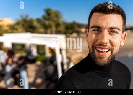 Positivo giovane uomo bearded in nero dolcevita con orecchini guardando la macchina fotografica mentre in piedi su sfondo sfocato della zona di campagna tropicale Foto Stock