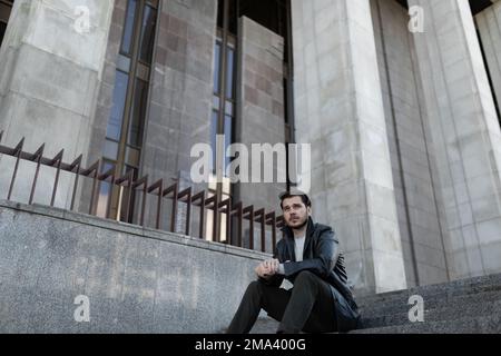 un uomo triste e sconvolto seduto sui gradini vicino all'edificio grigio Foto Stock