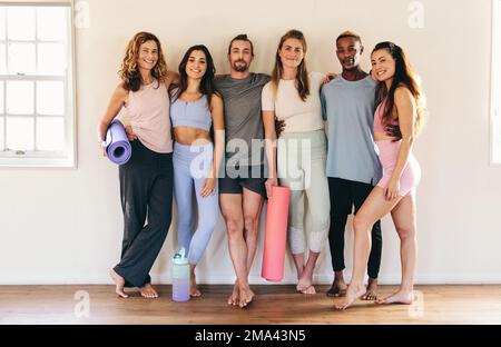 Ritratto degli amici di fitness che si abbracciano mentre si trovano in piedi in uno studio di yoga. Gruppo di persone diverse che frequentano una lezione di yoga insieme. Buon pisello Foto Stock