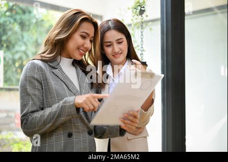 Due splendide donne d'affari o colleghi asiatici millennial discutono, condividono le loro idee commerciali, rivedono insieme il rapporto finanziario. Foto Stock