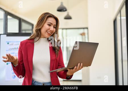 Una donna d'affari asiatica, professionista ed entusiasta, si trova nel suo ufficio utilizzando un computer portatile, soddisfatto dei risultati del progetto. Foto Stock