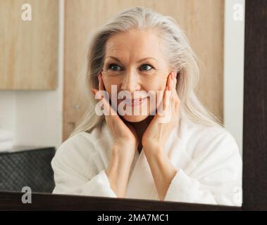 Ritratto di una donna matura sorridente con capelli grigi che guarda uno specchio in bagno Foto Stock
