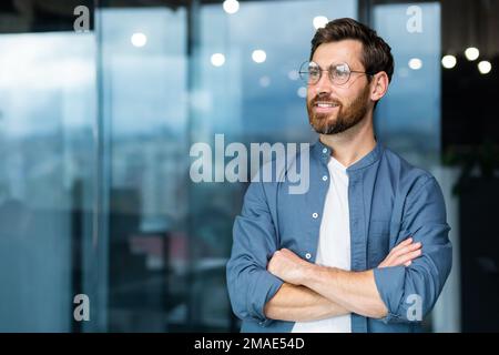 Ritratto di un giovane uomo di successo, uomo d'affari, freelance, designer in piedi in ufficio, incrociando le braccia, guardando con fiducia al lato Foto Stock