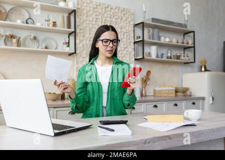 Giovane donna arrabbiata seduta in cucina a casa con un computer portatile. Contiene documenti, una lettera nelle mani, digita sul telefono. Foto Stock