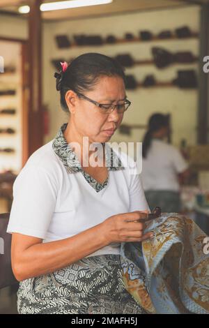 Bali - Indonesia - 10.22.2015: Anziana donna indonesiana che disegna batik su tessuto blu in un atelier turistico Foto Stock